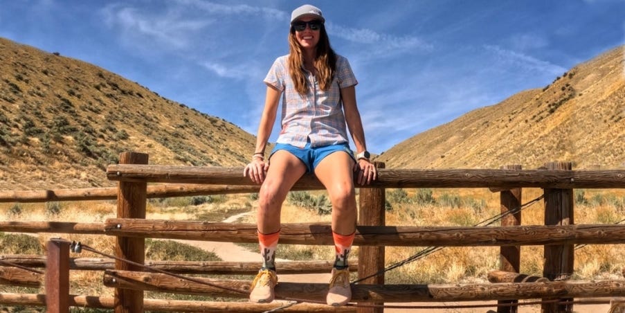 Trish Arends sitting on a wooden fence with a mountain trail in the background.