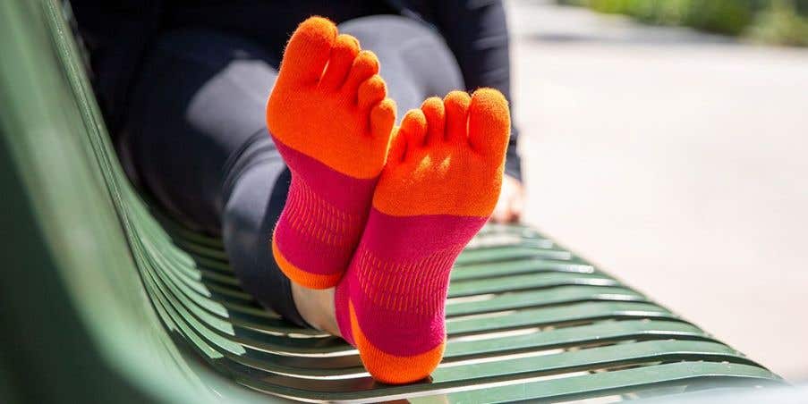A person sitting on a green bench with their feet up wearing Injinji Women's Run Lightweight No-Show socks in Chili.