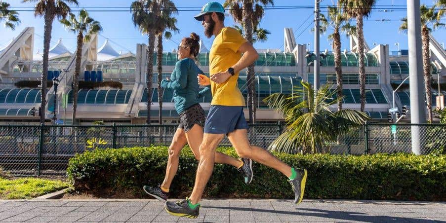 Two people running on a wide city sidewalk with palmtrees behind them.