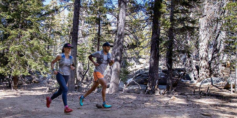 Two trail runners mid-stride as they run through a forested trail on a sunny day.