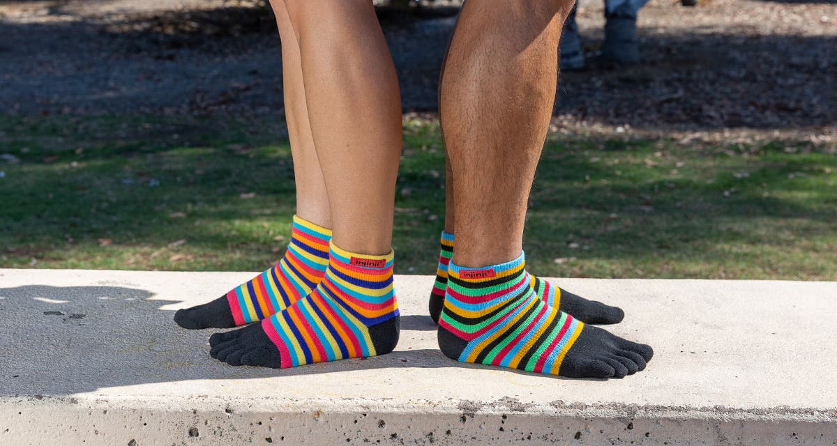 Two people standing back to back on a concrete bench in a park wearing Injinji Sport Original Weight Mini-Crew Toesocks in Rainbow and Sunset colorways.  