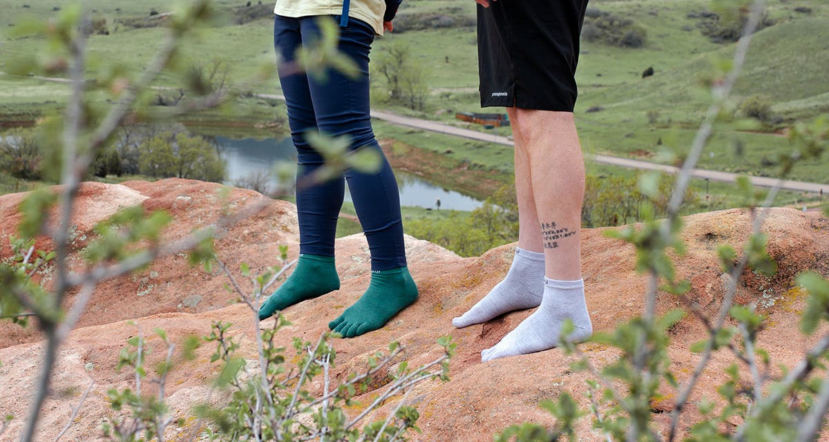 Two hikers wearing Injinji Liner Mini-Crew toesocks standing on a rock with a lake and a trail in the background
