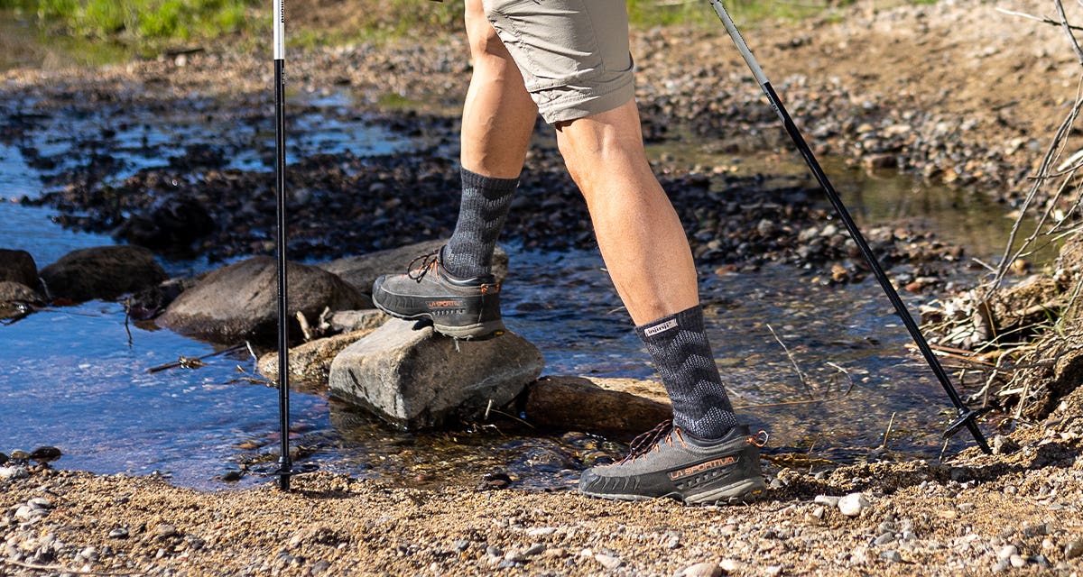 A man wearing hiking apparel and Injinji Outdoor Midweight Crew Wool performance socks crossing a creek using hiking poles. 