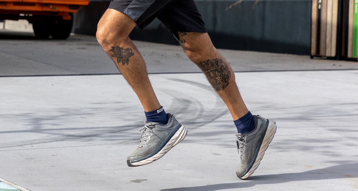 A man in athletic clothing running on a wide sidewalk wearing navy Injinji Run Original Weight Mini-Crew performance socks and running shoes. 