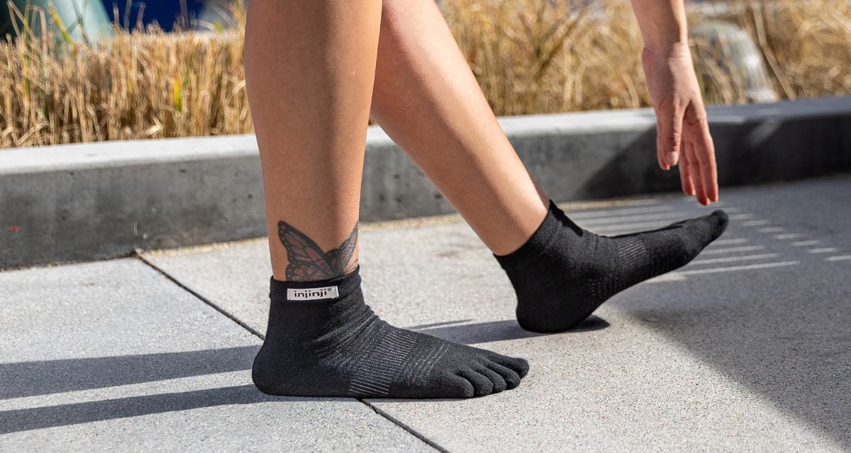 A close-up of a woman's bare calves and feet wearing Injinji Run Original Weight Mini-Crew black unisex running socks, reaching her fingers to her toes. 