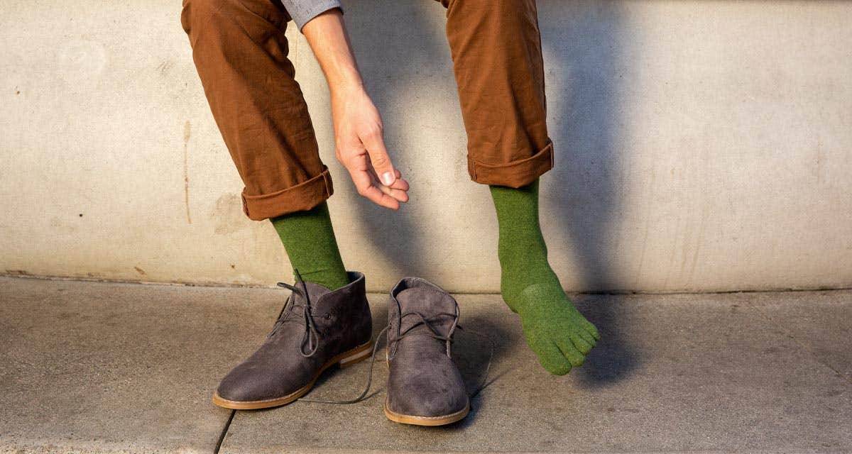 A man wearing dress pants and dress shoes leaning against a wall reaching to adjust his Injinji Everyday Crew Upcycled Cotton toesocks in Musgo. 