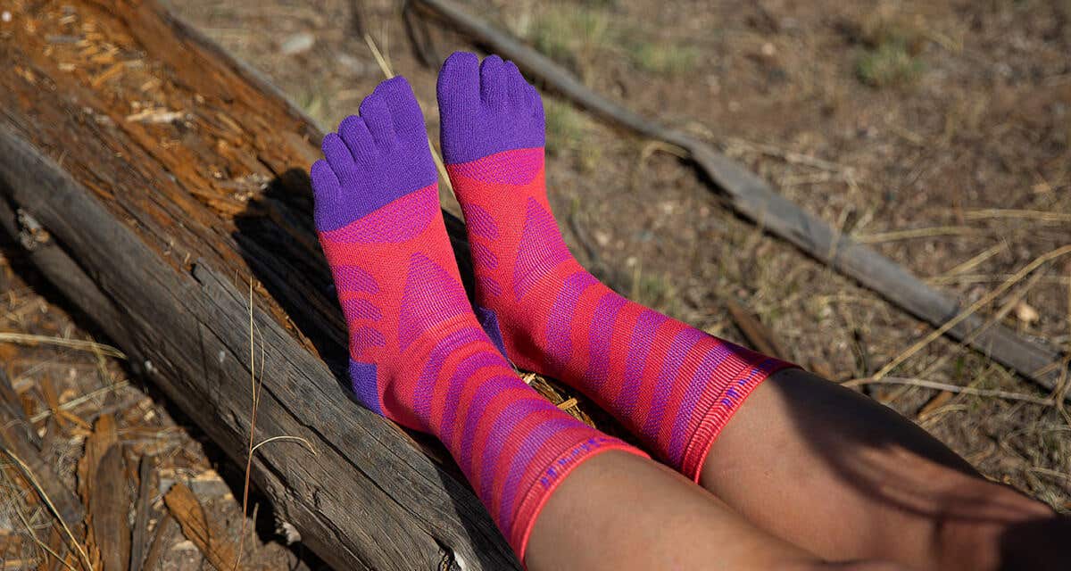 A woman sitting on a downed log with her legs outstretched wearing Injinji Women's Ultra Run Mini-Crew toesocks in Razzmatazz.