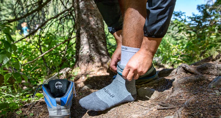 A man standing on a wooded trail adjusting his Injinji Liner + Hiker Crew performance toesocks. 