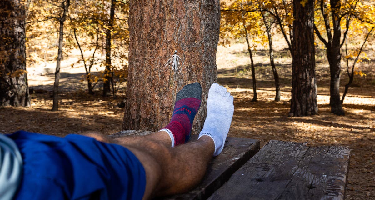 A man sitting down with his legs outstretched on top of a picnic table in a wooded campground wearing the Injinji Men's Liner + Runner Mini-Crew in brick. 