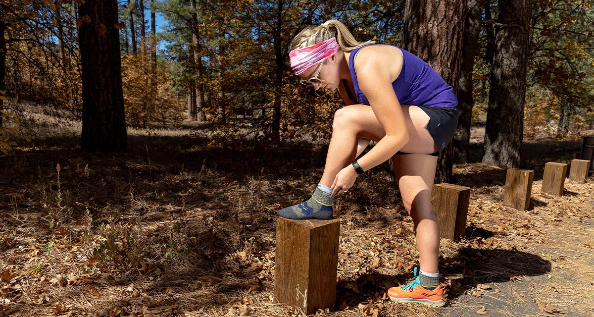 A woman in hiking apparel stopped on a trail resting her foot on a wooden stump to adjust her Injinji Woman's Liner + Runner Mini-Crew. 