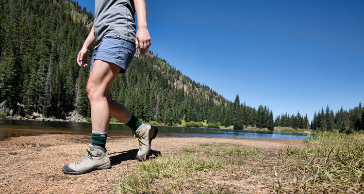 A woman wearing hiking apparel walking next to an alpine lake in hiking boots and Injinji Women's Hiker + Liner Crew performance toesocks