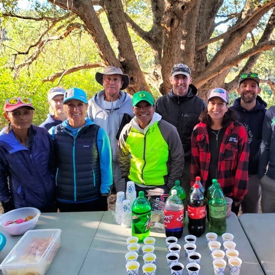 Eight member of the "San Diego Ultra Running Friends" club standing outdoors behind the table with drinks