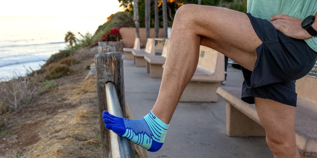 A person using a beach side barrier fence to stretch their leg while wearing Injinji Ultra Run Mini-Crew socks.