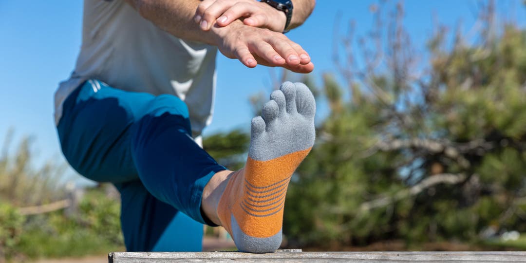 A closeup of a man stretching his hamstrings using a park bench as a prop to hold his leg up as he stretches his hand to his toes. He is wearing Injinji Run Lightweight No-Show toesocks in orange and gray, plus blue track pants and a grey t-shirt. 