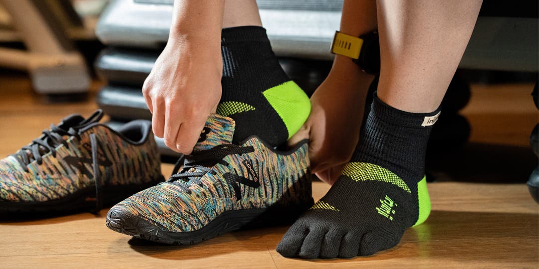 A closeup shot of a woman wearing the Injinji Trainer Mini-Crew lime green and black gym sock, as she puts on her black and multicolored New Balance gym shoes. 