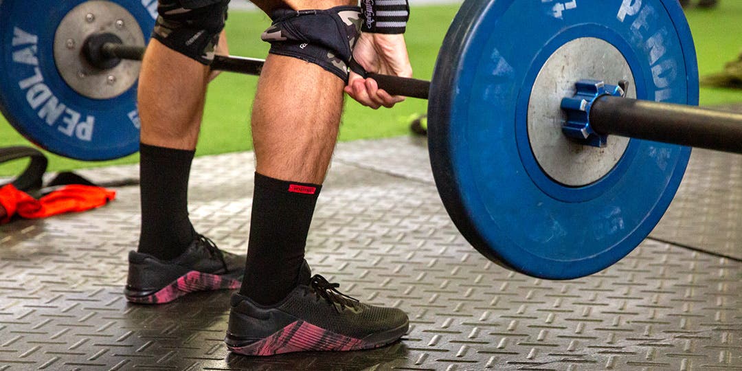 A closeup of the male weightlifters black trainers, black Injinji Sport Original Weight Crew toesocks, calves, and knees, performing a deadlift with a large dumbbell. 
