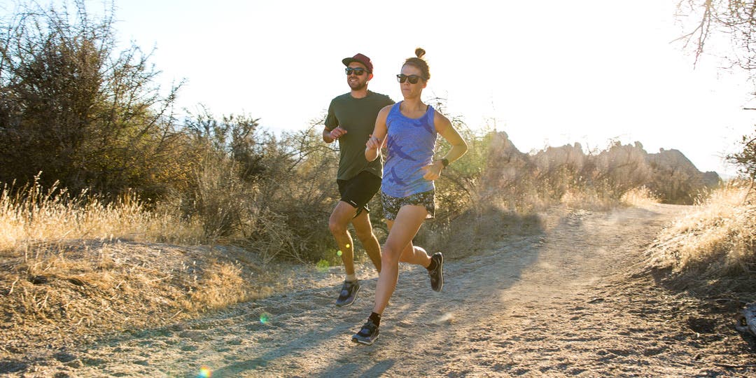 A man and woman running side by side on a sandy trail surrounded by desert shrubs. She's wearing a blue tank top, running shorts, running shoes and Injinji toesocks. He's wearing a black hat, black shorts, grey t -shirt, running shoes with toesocks..