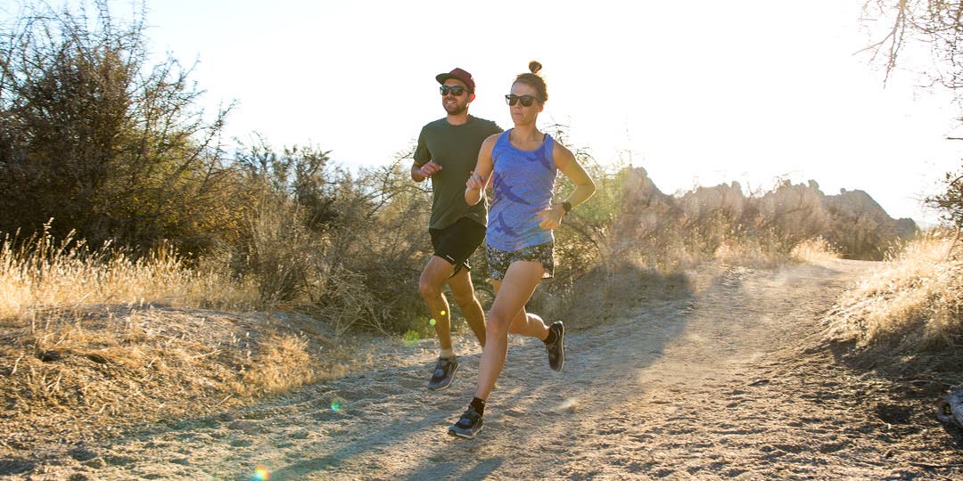 Three pepole running on a sandy, mostly flat trail among desert shrubs.