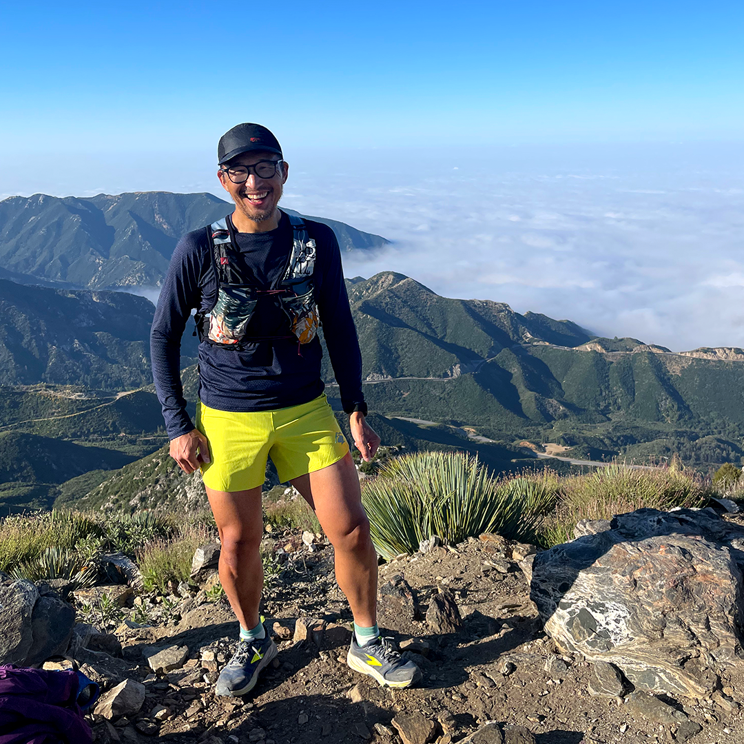 Team Injinji athlete Phil Shin resting on top of a mountain with clouds below him in the background.