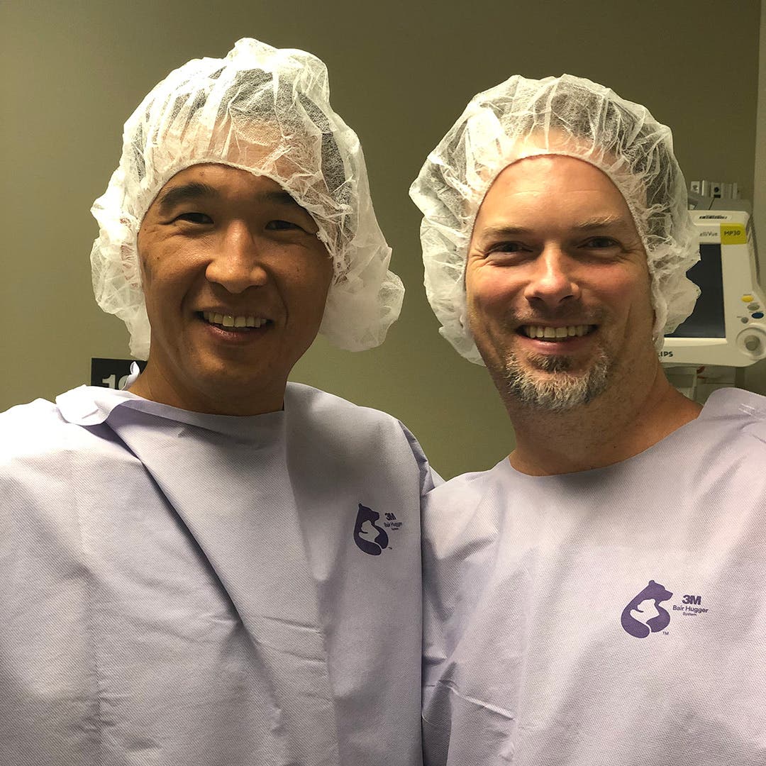 Team Injinji athlete Phil Shin standing in the hospital with his live liver transplant doner Murph both wearing hospital gowns and hair nets.