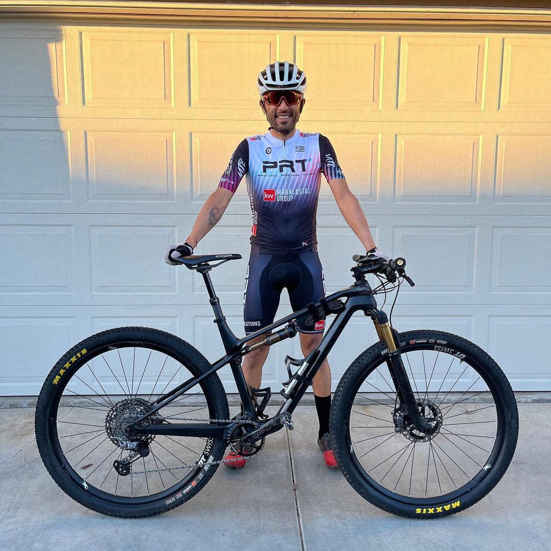 Team Injinji Athlete Jesus Topete Jr. outfitted in bike racing gear, holding up his bike while standing in front of his garage door. 