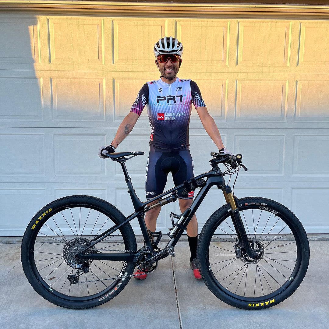 Team Injinji Athlete Jesus Topete Jr. outfitted in bike racing gear, holding up his bike while standing in front of his garage door.