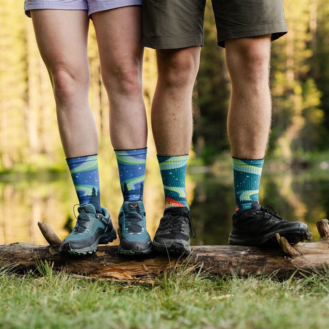 Two people standing on a log in front of a lake wearing Injinji Artist Designed toesocks in Howl and Camp.