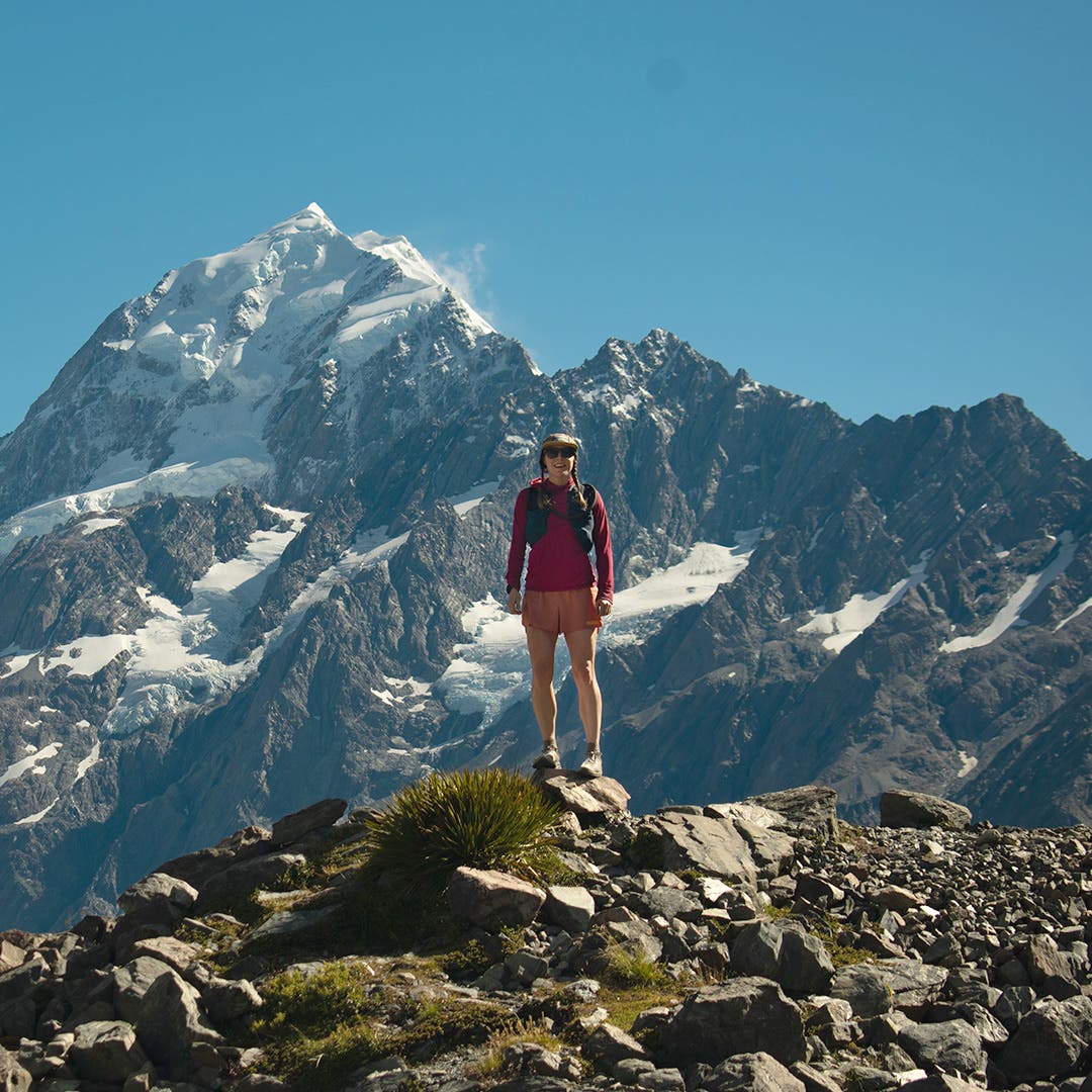 Team Injinji Athlete Caroline Himbert standing on some rocks at the edge of a mountain with epic views of mountain peaks behind her.