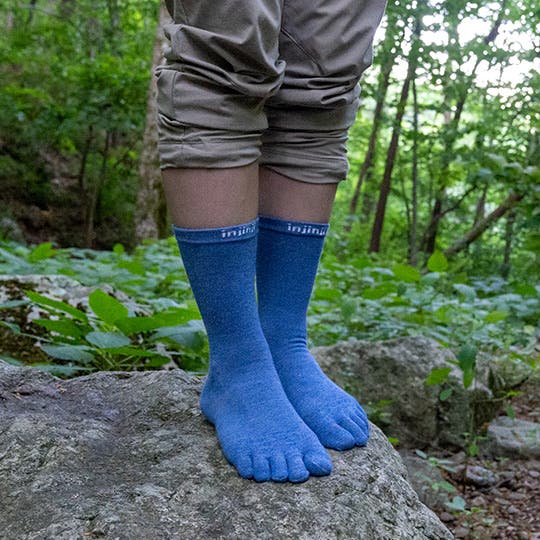 A hiker standing on top of a rock next to a lush green trail, wearing blue Injinji Liner Crew toesocks.