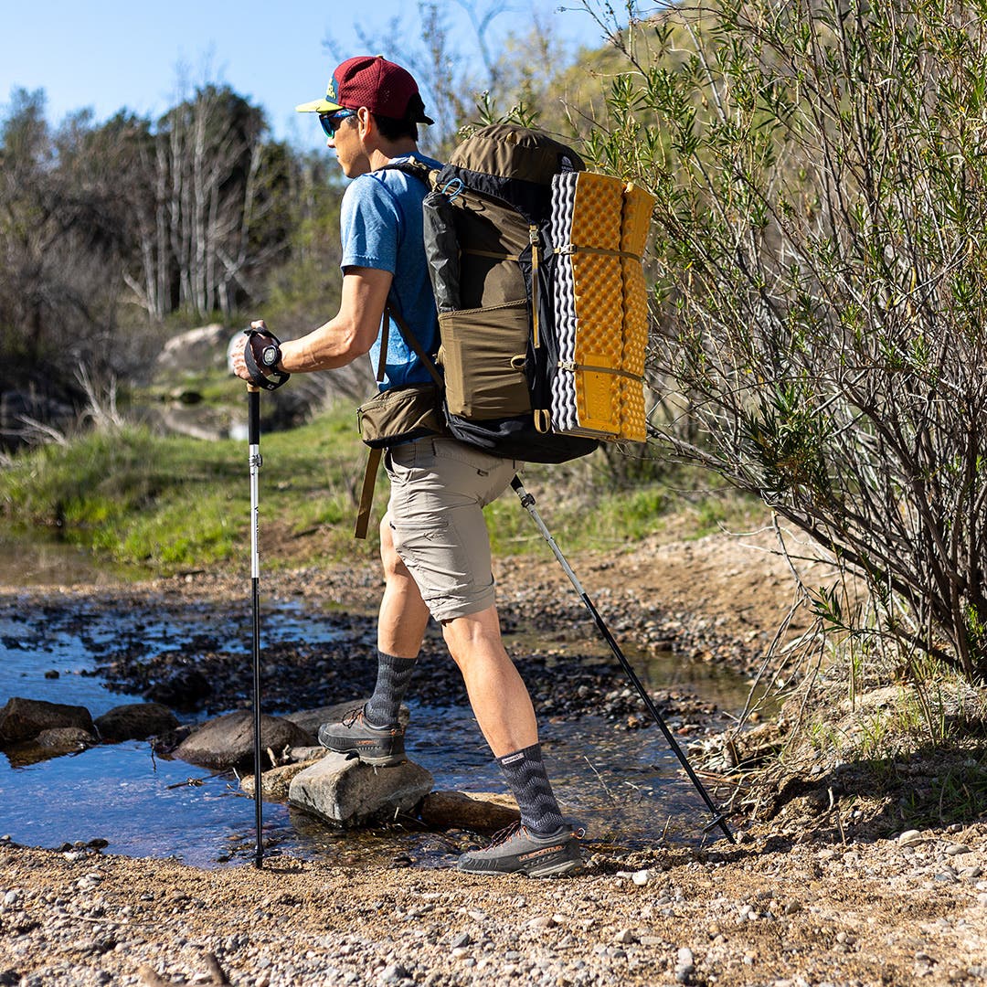 Backpacker with a pack and poles crossing a creek wearing hiking shoes and Injinji Outdoor Midweight Crew wool socks. 