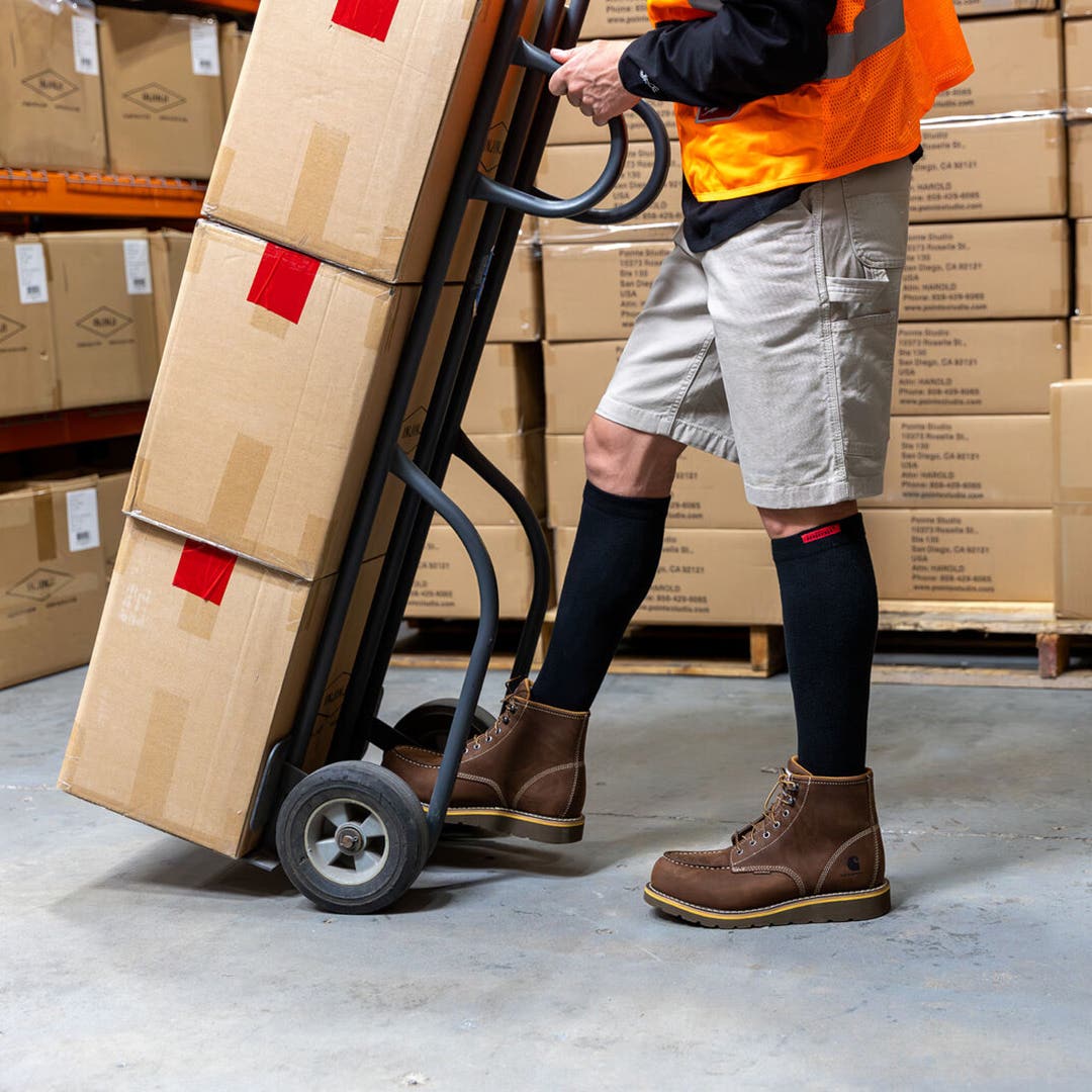 A person using a dolly piled with cardboard boxes in a warehouse wearing Injinji Boot Original Weight OTC socks underneath their work boots.