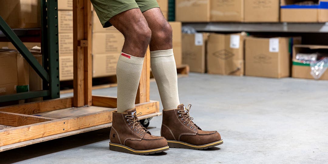 A person leaning against a wooden work bench in a warehouse wearing Injinji Boot Original Weight OTC socks underneath their work boots.