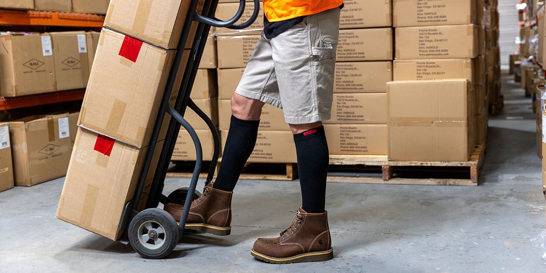 A person using a dolly piled with cardboard boxes in a warehouse wearing Injinji Boot Original Weight OTC socks underneath their work boots.