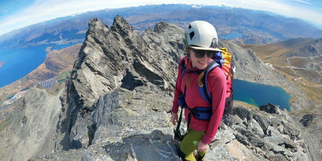 Team Injinji Athlete Caroline Himbert standing in her climbing gear at the top of a jagged mountain with alpine lakes in the background.