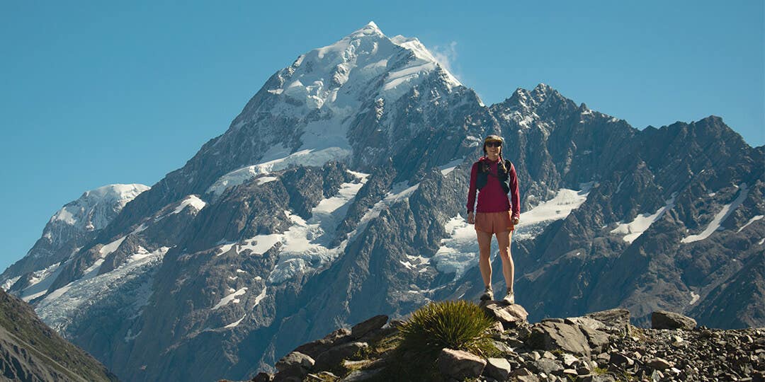 Team Injinji Athlete Caroline Himbert standing on some rocks at the edge of a mountain with epic views of mountain peaks behind her.