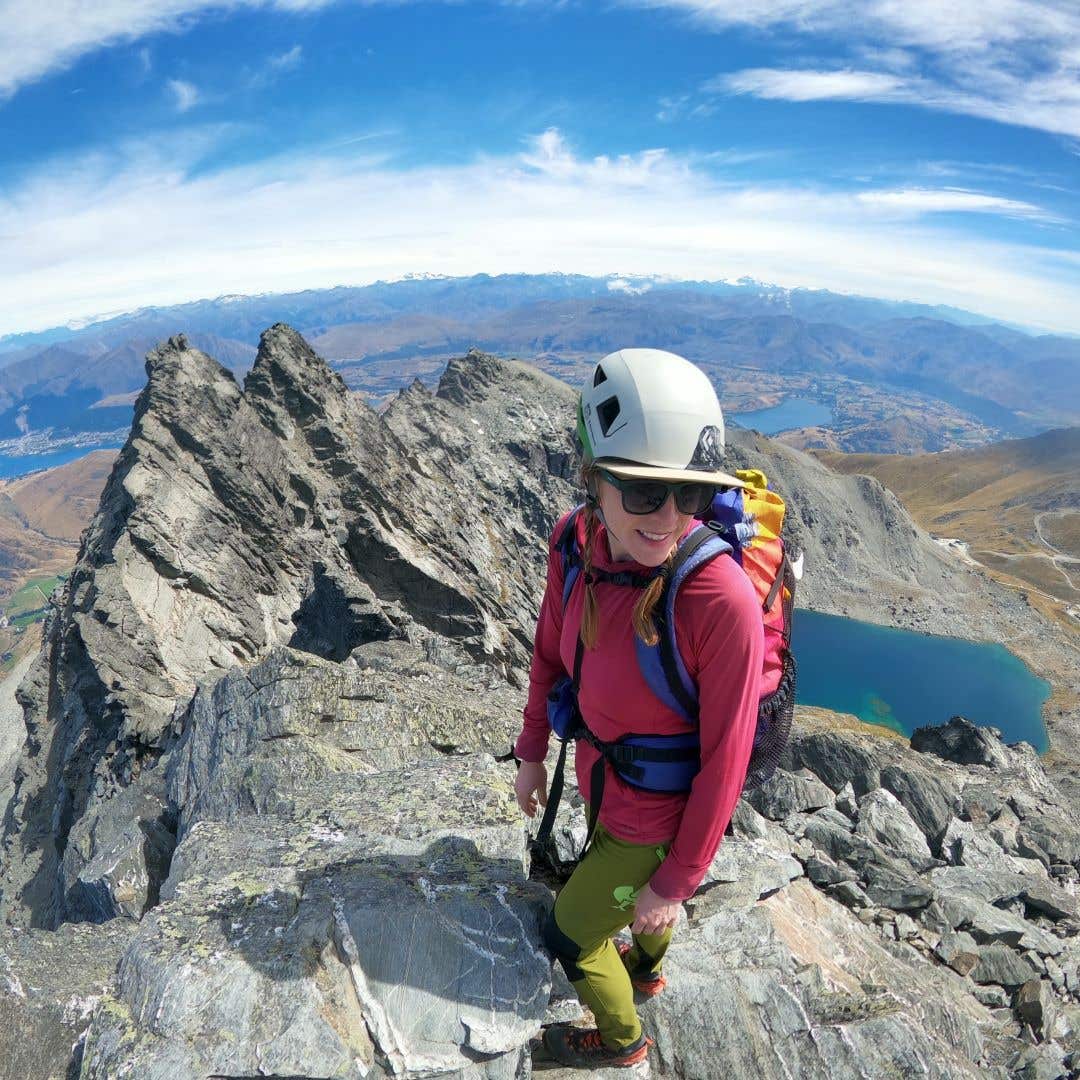 Team Injinji Athlete Caroline Himbert standing in her climbing gear at the top of a jagged mountain with alpine lakes in the background.