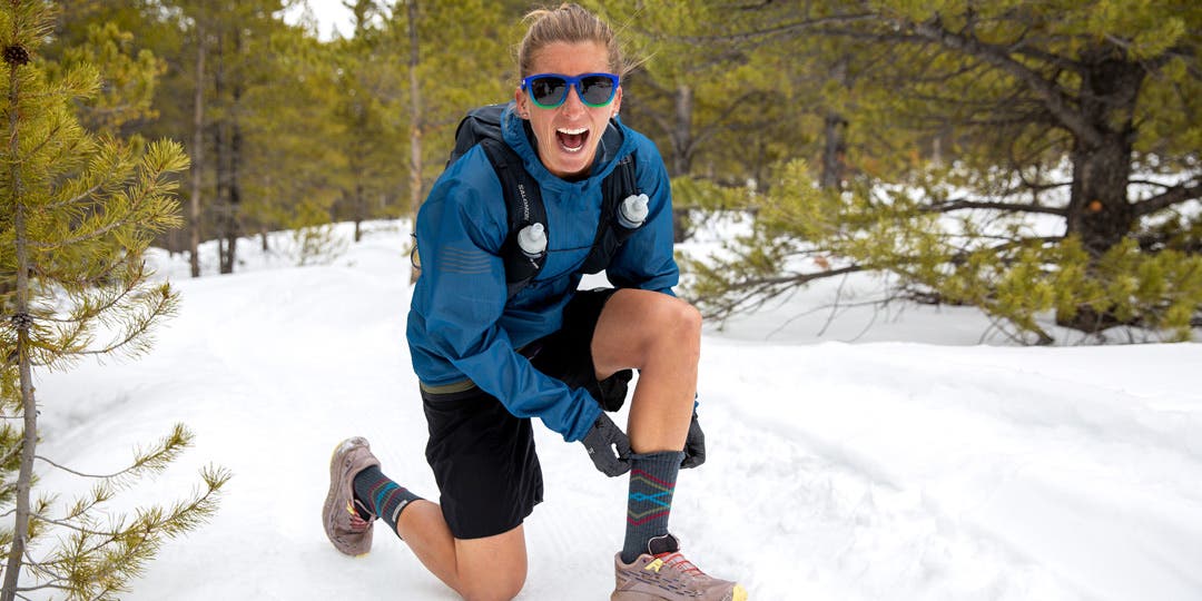 Courtney Dauwalter in a snowy forest with her knee down to adjust her Injinji Trail Midweight Crew sock.