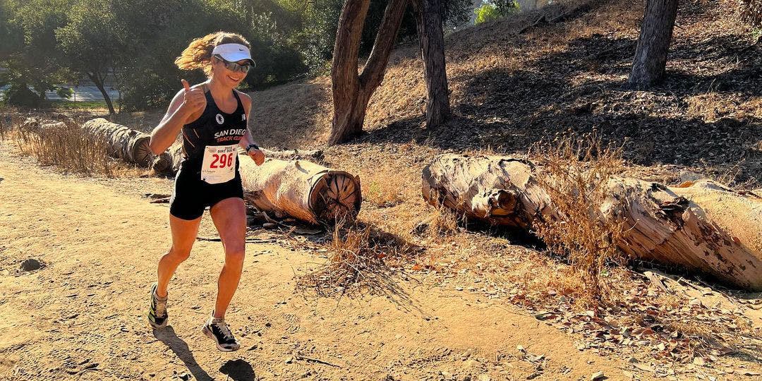 A female running on a dirt trail, wearing a black tank top and shorts, with downed trees in the background on a sunny day.