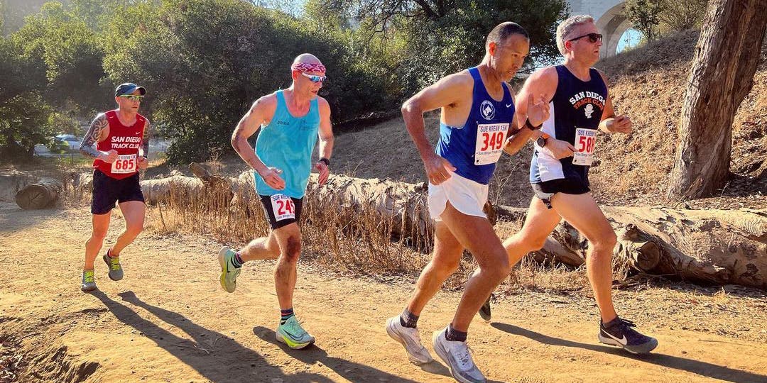 A group of four middle aged, males wearing multicolored shorts and tanktops, running on a dirt trail with downed trees in the background.