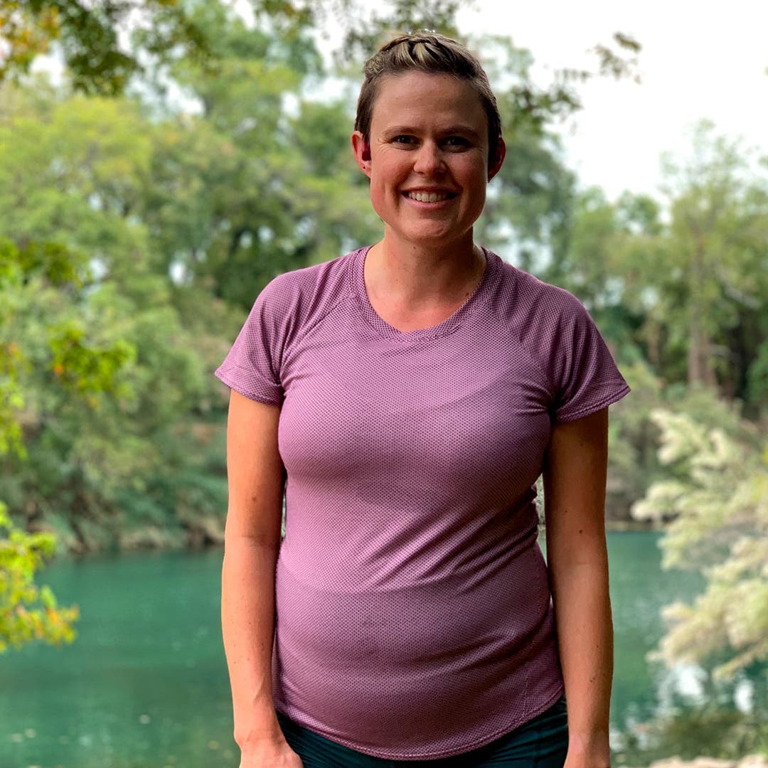 Cate Barrett pregnant and smiling in front of a lake and green foliage.