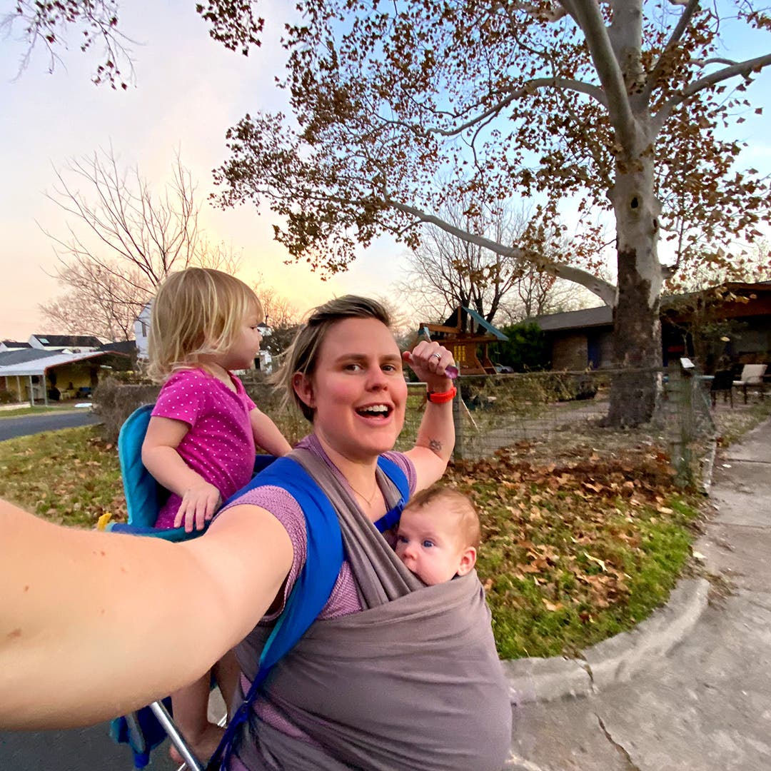 Cate Barrett on a walk carrying her newborn in a sling on her front side with her first child on her back.