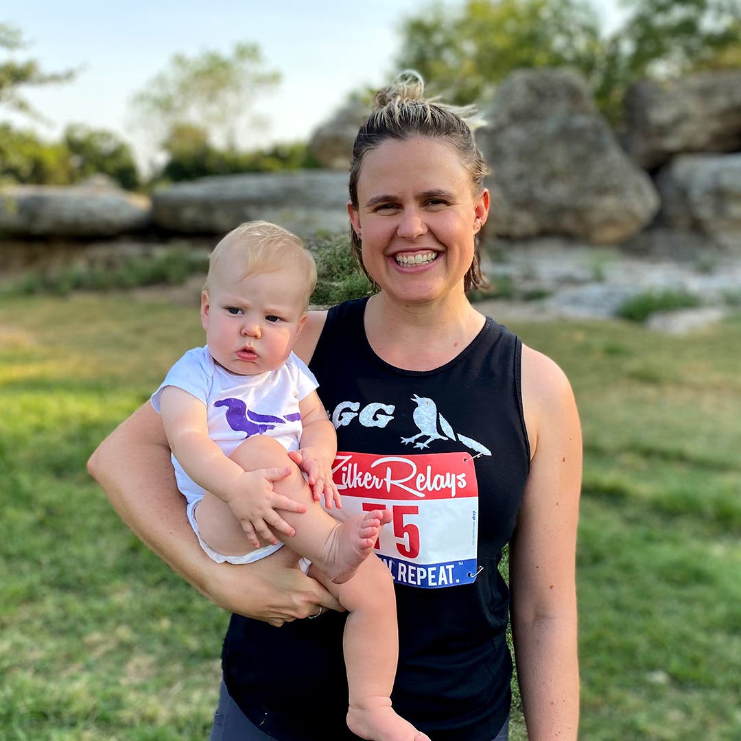 Cate Barrett smiling for the camera wearing a race bib and holding her infant.