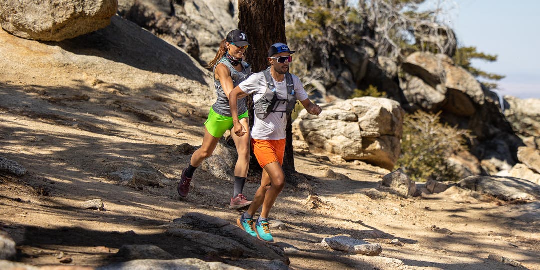 A man and woman running downhill on a mountainous trail.