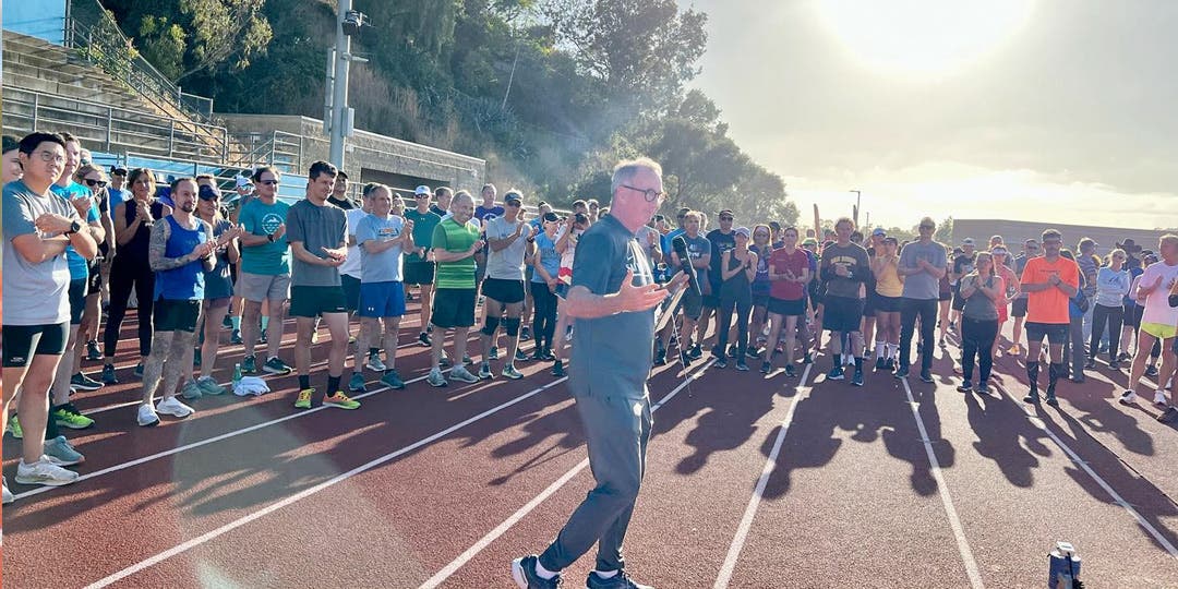 San Diego Track Club's Coach Paul Greer making a speech to a large group of athletes outside on a track.