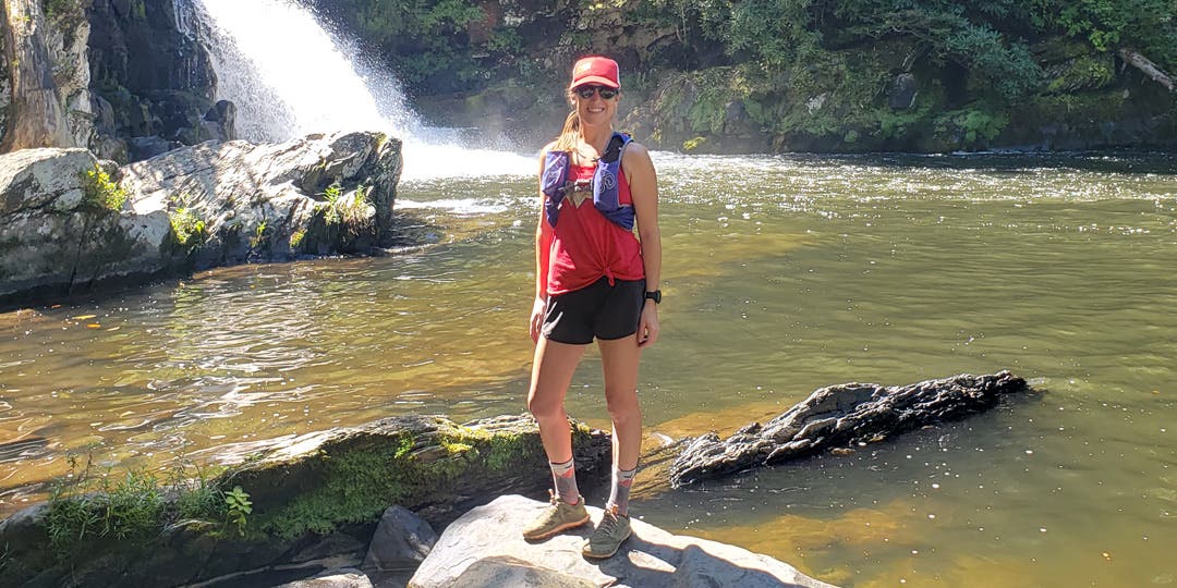 Female athlete wearing athletic apparel, a hat, and a hydration vest, standing in front of waterfall. 