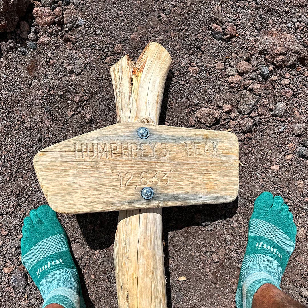 Both feet wearing Injinji Trail Midweight Mini-Crew socks straddling the sides of a Humphrey's Peak Trail sign.