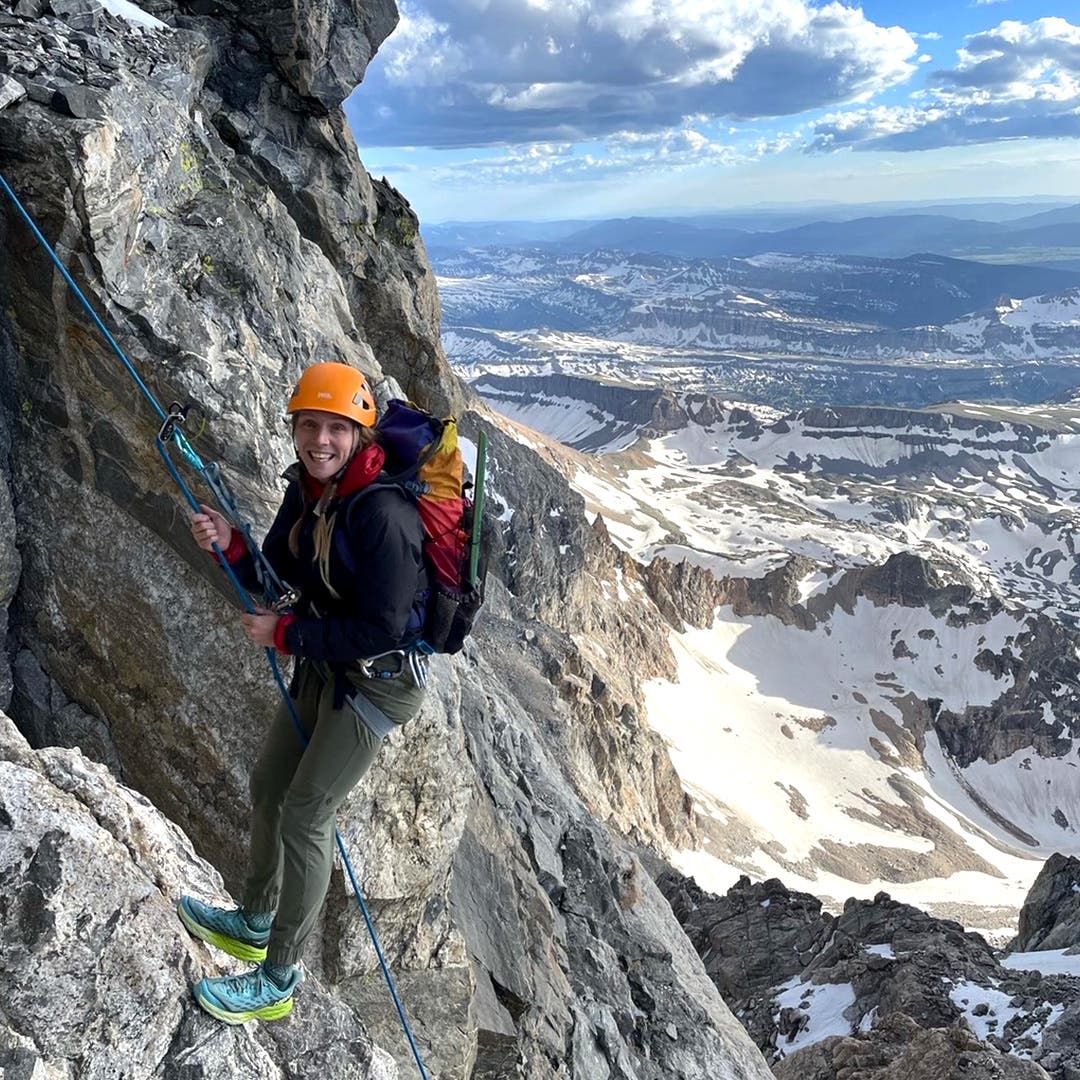 Team Injinji Athlete Caroline Himbert rock climbing an expansive view of snowy mountains behind her.
