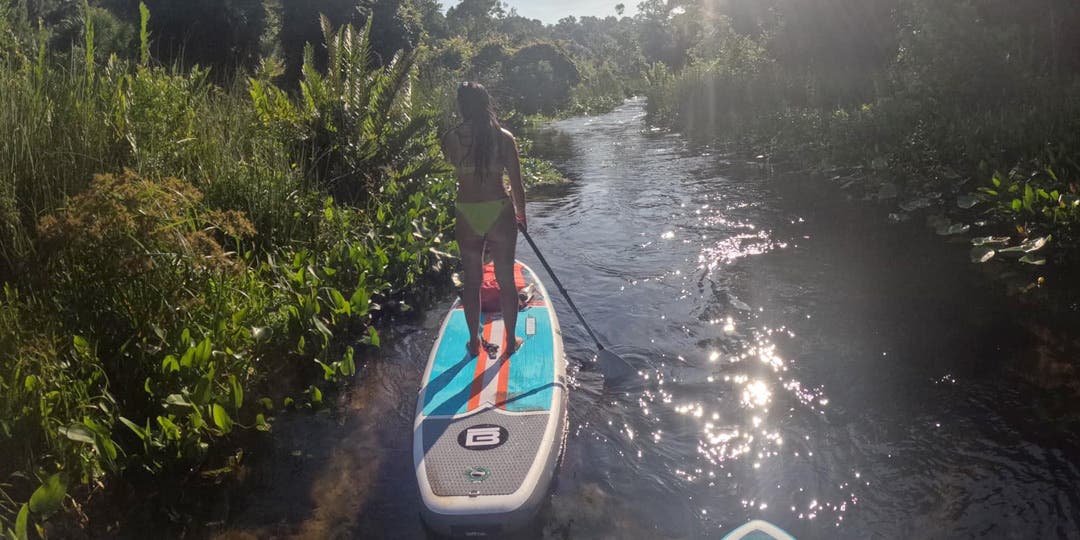 Team Injinji Athlete Lovelyn Findley standing on a paddle board paddling down a river surrounded by lush green foliage.