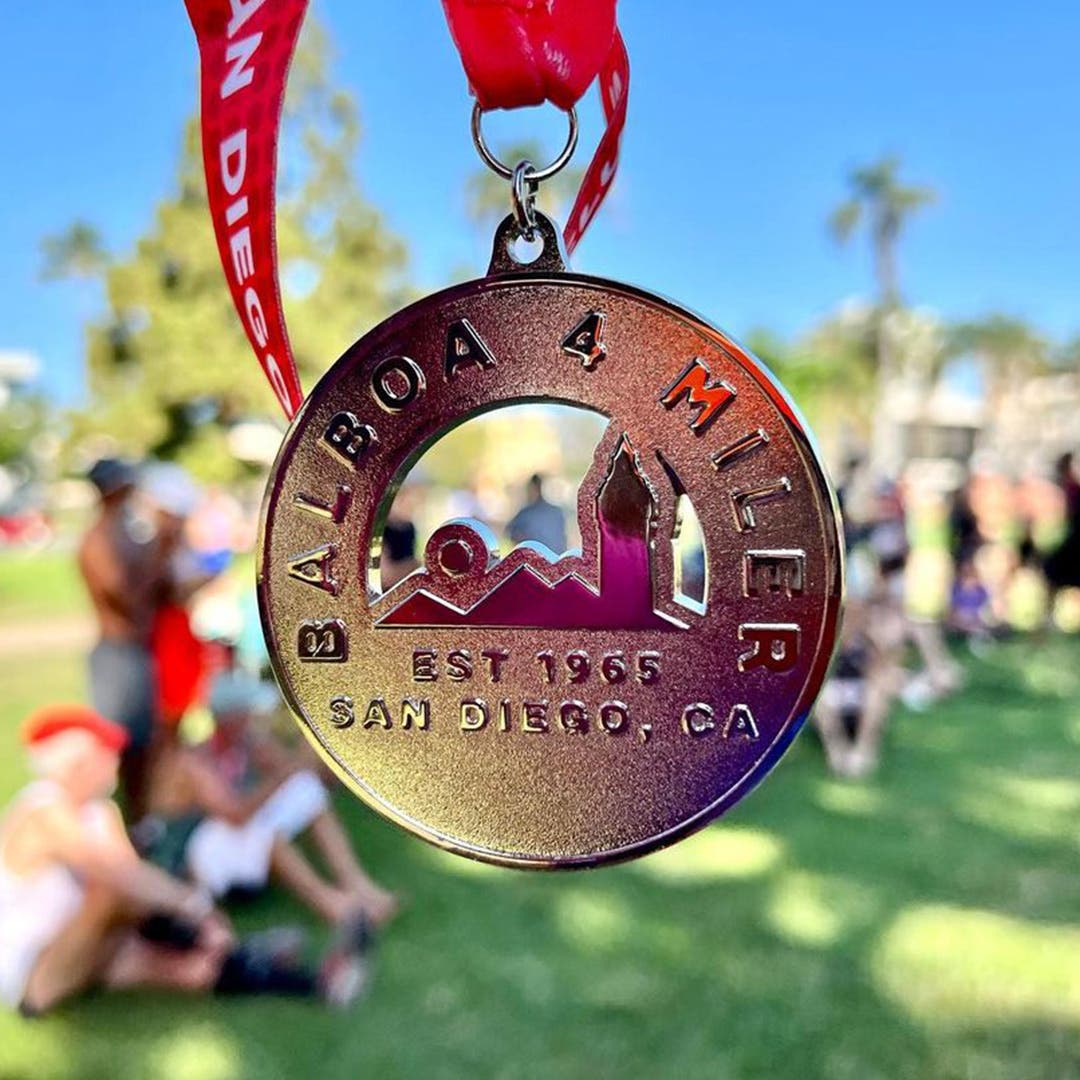 A closeup of a gold medal given to the winners of the Balboa 4 Miler race in San Diego, with people and green grass out of focus in the background. 