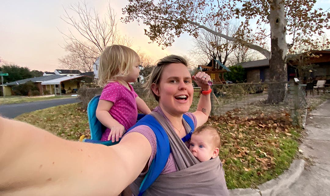 Cate Barrett on a walk carrying her newborn in a sling on her front side with her first child on her back.